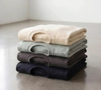Four folded blank apparel garments in neutral colors, arranged from cream to navy on a concrete floor.