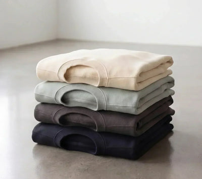 Four folded blank apparel garments in neutral colors, arranged from cream to navy on a concrete floor.