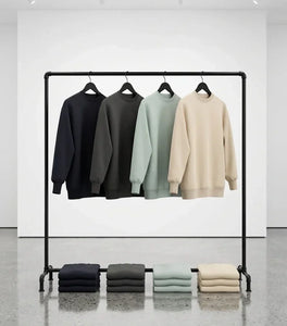 Four sweatshirts displayed on a black pipe rack with folded sweatshirts underneath in navy, charcoal, sage, and cream.