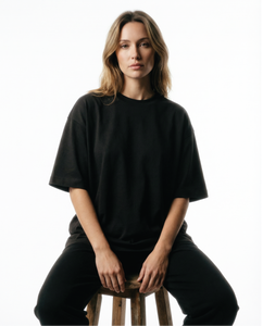 Woman wearing a black Basico Branco oversized organic cotton T-shirt while seated on a stool in a minimalist studio.