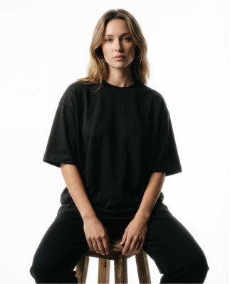 Woman wearing a black oversized tee by Basico Branco sitting on stool against clean studio background.