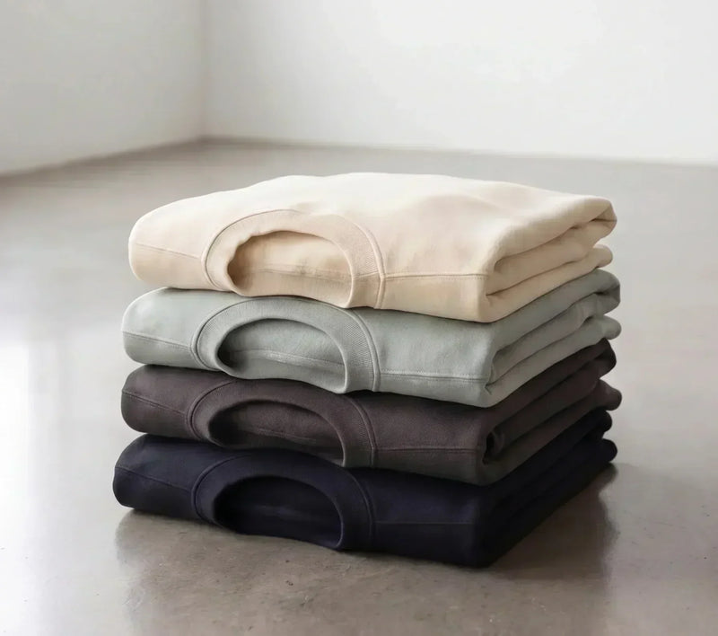 Four folded blank apparel garments in neutral colors, arranged from cream to navy on a concrete floor.