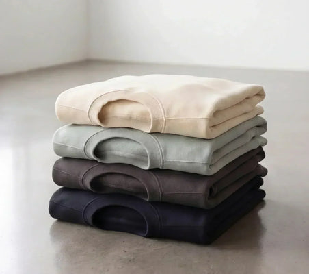 Four folded blank apparel garments in neutral colors, arranged from cream to navy on a concrete floor.