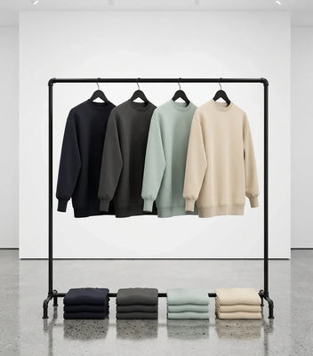 Four sweatshirts in black, charcoal, sage, and cream displayed on a metal rack with folded versions below.