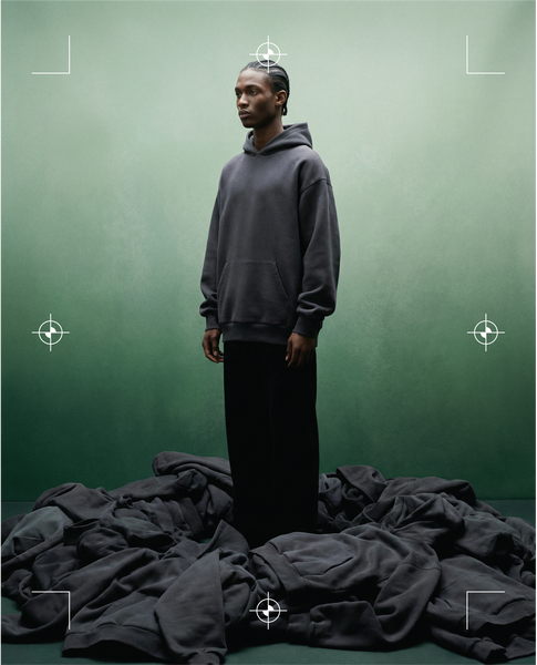 Basico Branco charcoal hoodie worn against a green backdrop with stacked apparel below.