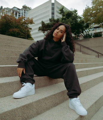 Woman in a black sweatsuit and white sneakers sitting on outdoor steps in an urban setting.
